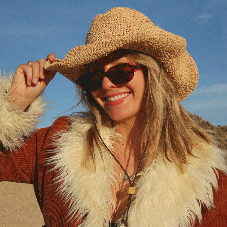 Woman wearing a straw hat, sunglasses, and a fur-trimmed coat against a blue sky.