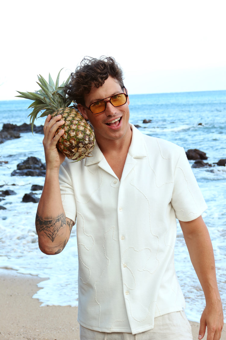 A man wearing a white short-sleeve button-up shirt and light-colored pants stands on a beach holding a pineapple to his ear. He is wearing yellow-tinted sunglasses, with the ocean and rocks in the background.