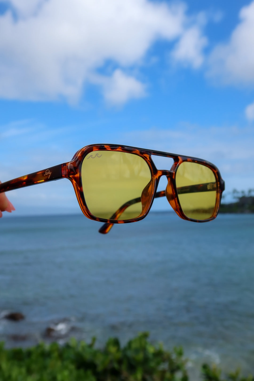 A pair of square aviator-style sunglasses with a tortoiseshell frame and yellow tinted lenses, held up by a person's hand against a blurred ocean and sky background.