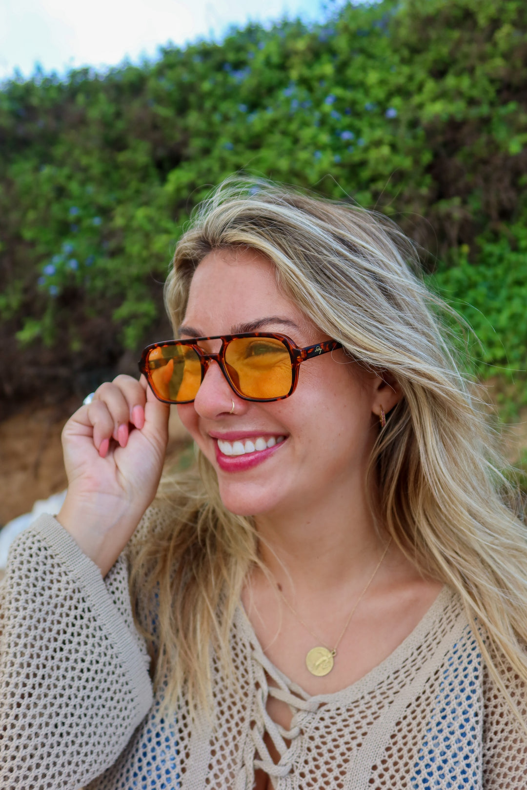A smiling blonde woman outdoors wearing tortoiseshell sunglasses with yellow-orange tinted lenses and a beige crochet top, with a blurred green hill in the background.