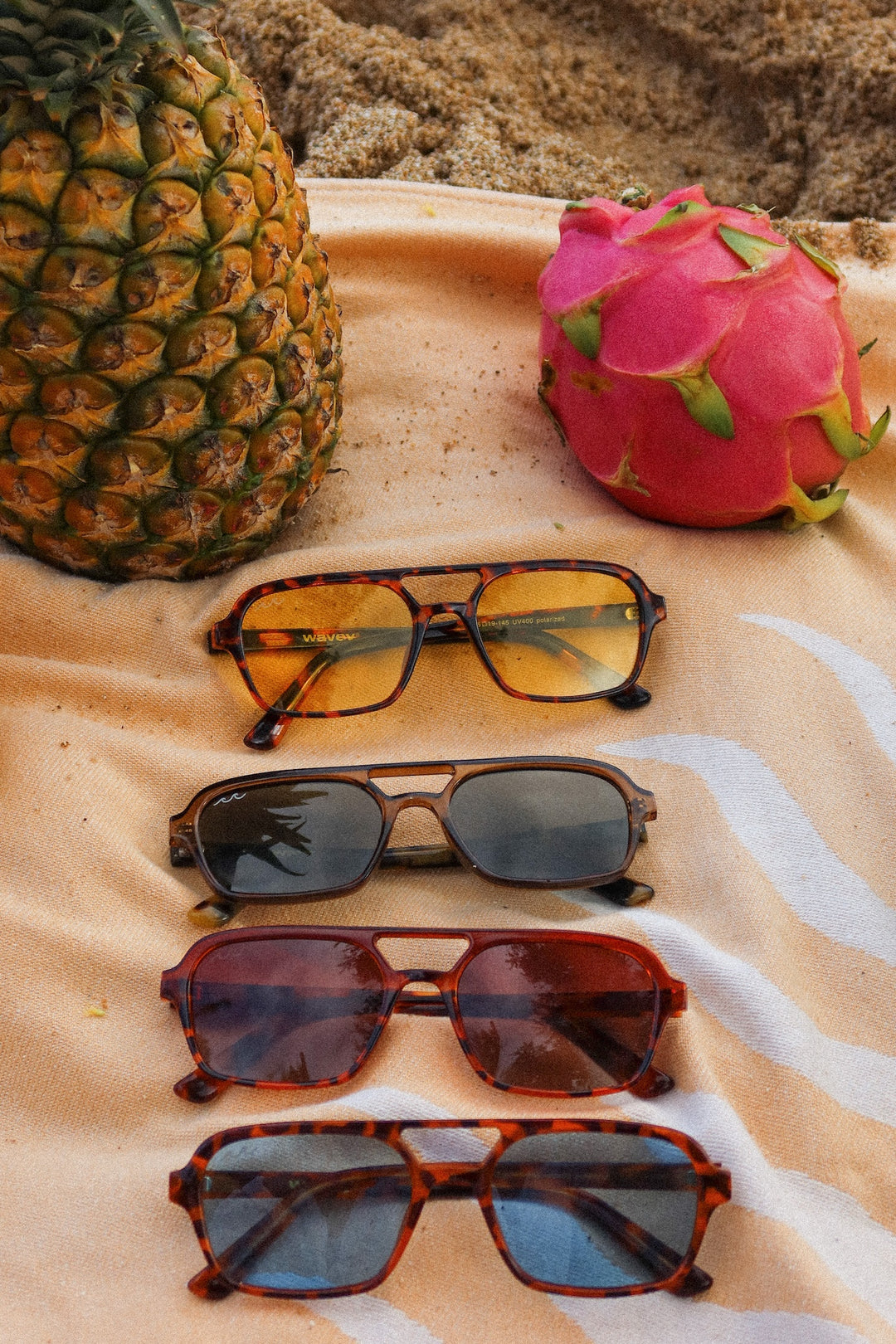 Four pairs of aviator-style sunglasses with tortoiseshell and dark lenses are arranged vertically on a beige towel, with a pineapple and a dragon fruit nearby on a sandy beach background.