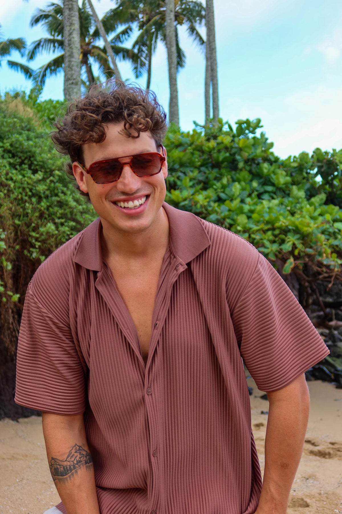 A smiling man wearing a short-sleeve, ribbed mauve-brown button-up shirt and sunglasses, standing outdoors on a sandy beach with palm trees and green foliage in the background.