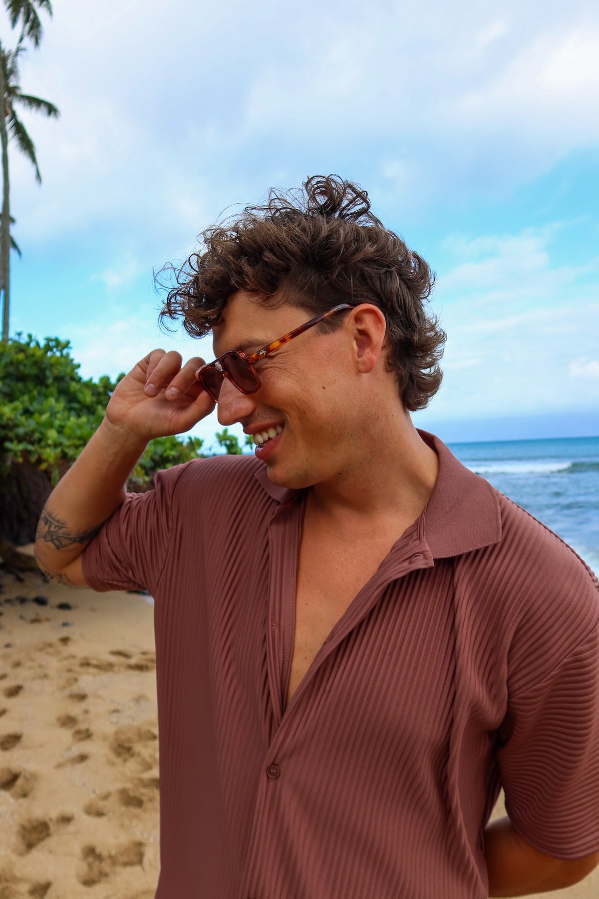 A man with curly brown hair smiling on a beach, wearing a brown pleated short-sleeve button-up shirt and tortoiseshell sunglasses.