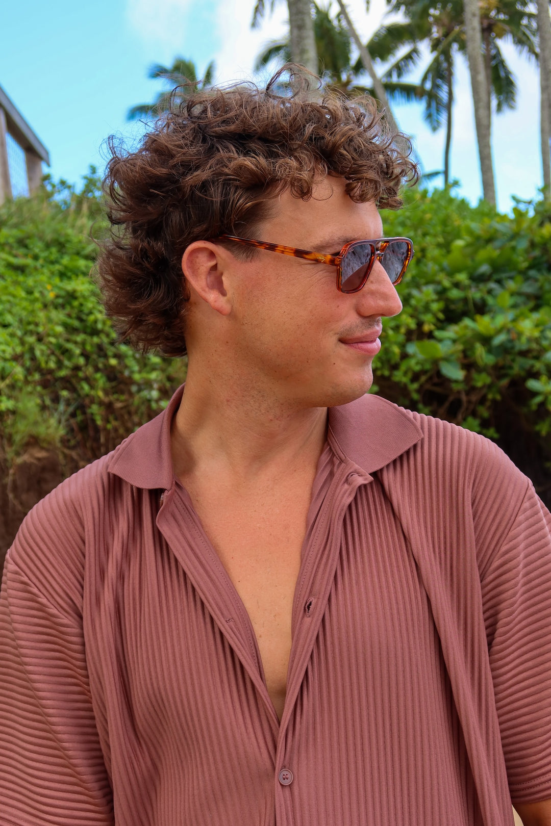 A man with curly brown hair wearing tortoiseshell sunglasses and a dusty rose pleated button-up shirt, photographed outdoors against a background of green foliage and palm trees.