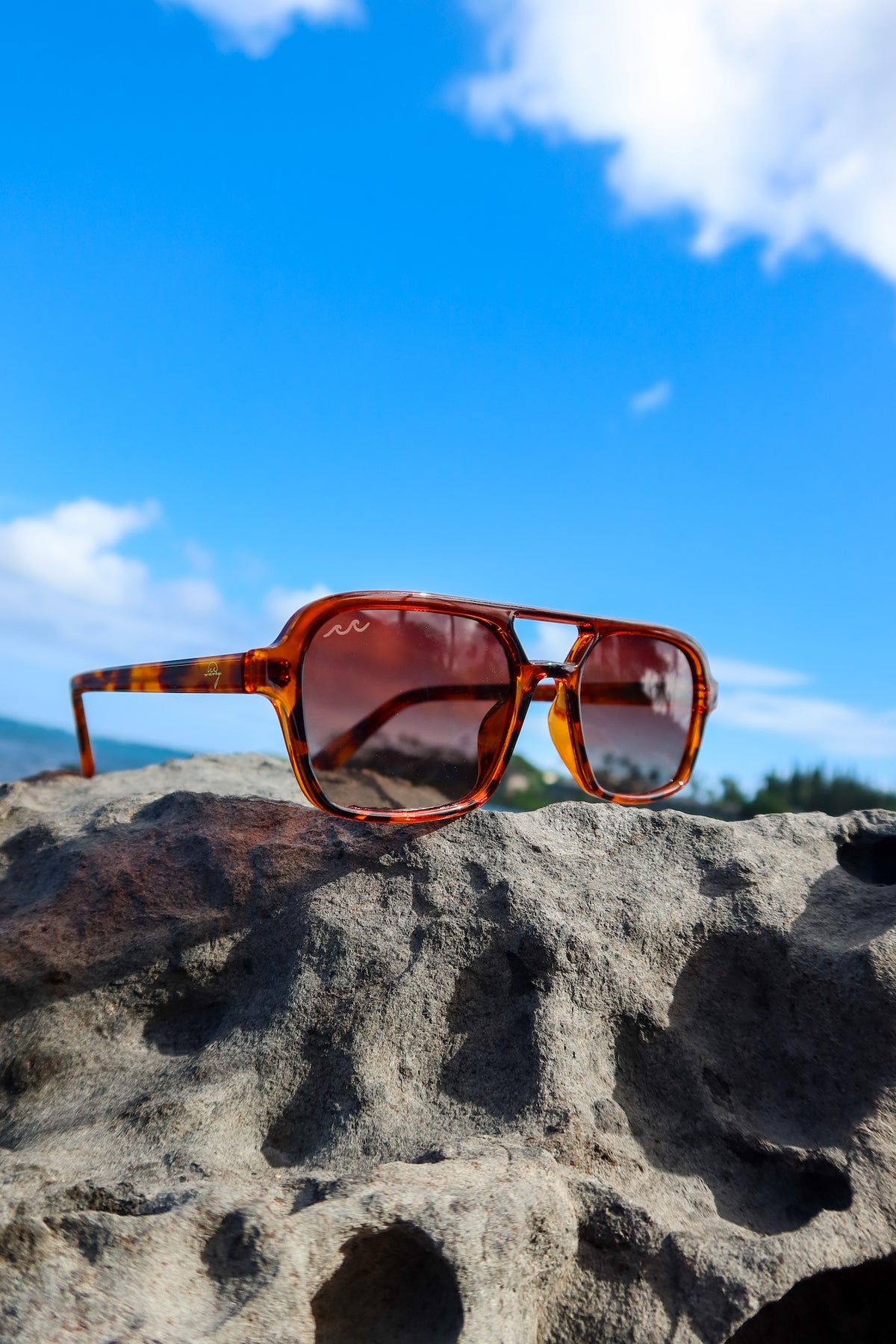 A pair of square sunglasses with a translucent amber-brown frame and dark tinted lenses, resting on a textured gray rock with a bright blue sky and clouds in the background.
