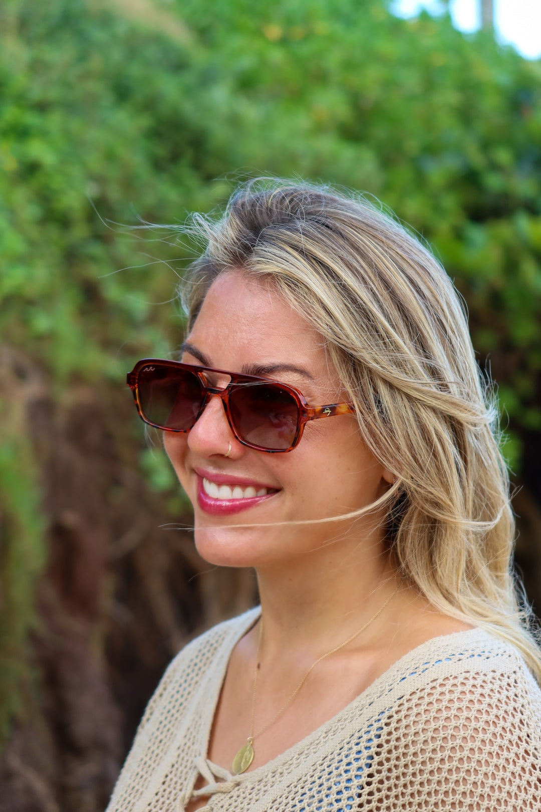 A woman with blonde hair wearing brown aviator-style sunglasses and a beige crochet top, photographed outdoors against a blurred green foliage background.