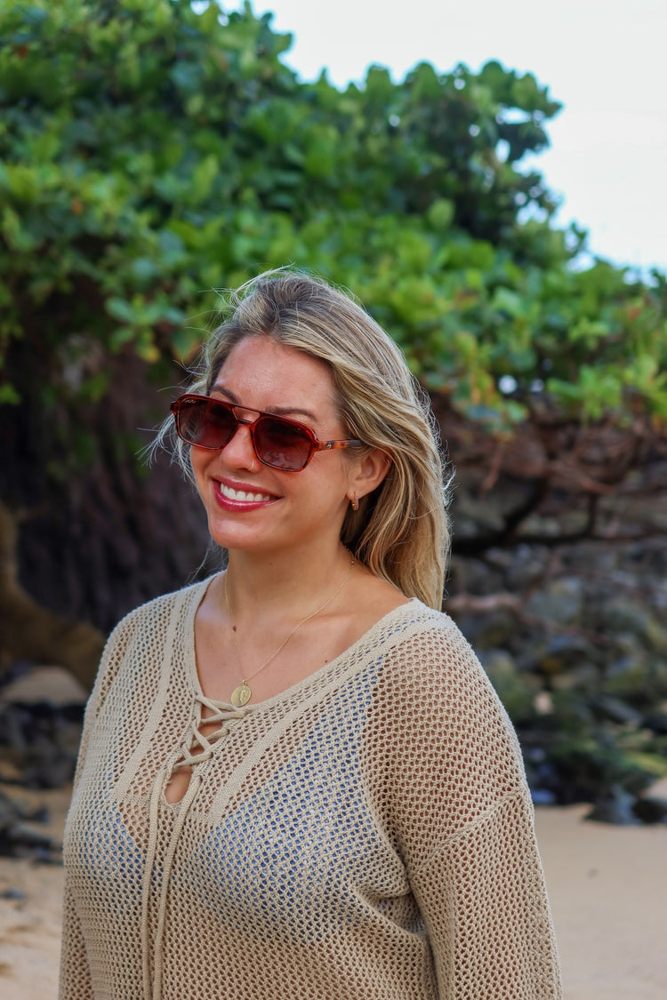 A smiling blonde woman wearing brown sunglasses and a beige open-knit lace-up top, standing outdoors against a background of green foliage and rocks.