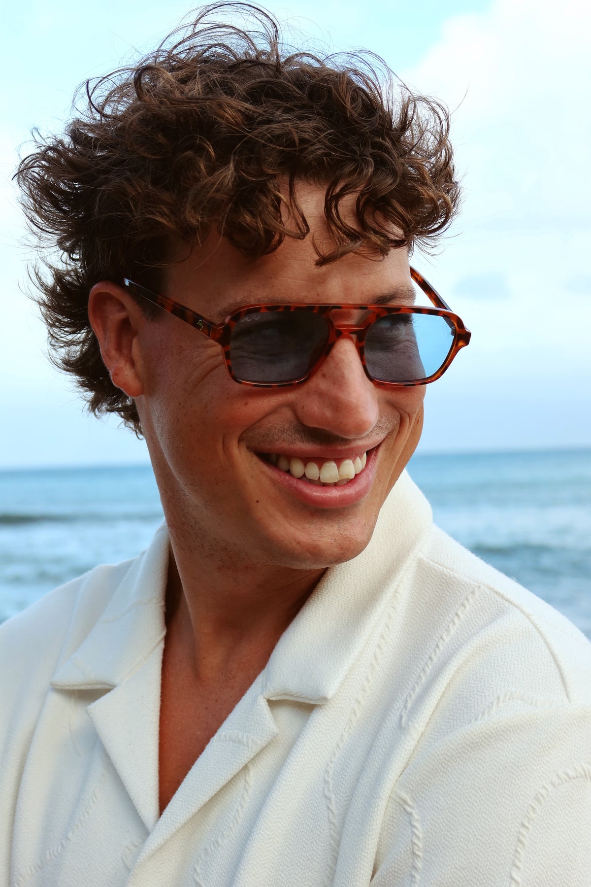 A smiling man with curly brown hair wearing tortoiseshell sunglasses and a white collared shirt, photographed outdoors with the ocean and sky in the background.