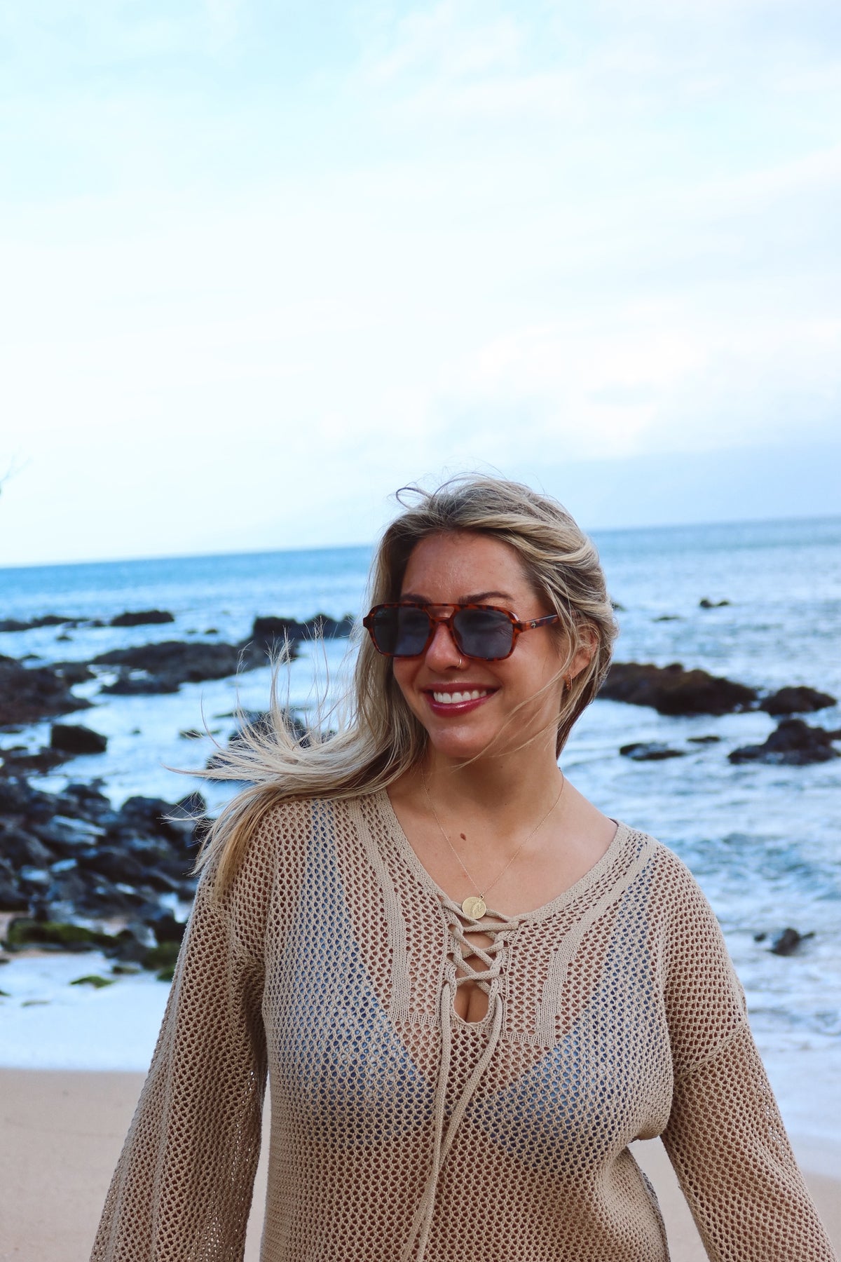 A woman with blonde hair and sunglasses stands on a beach wearing a beige open-knit long-sleeve top with lace-up front detailing.