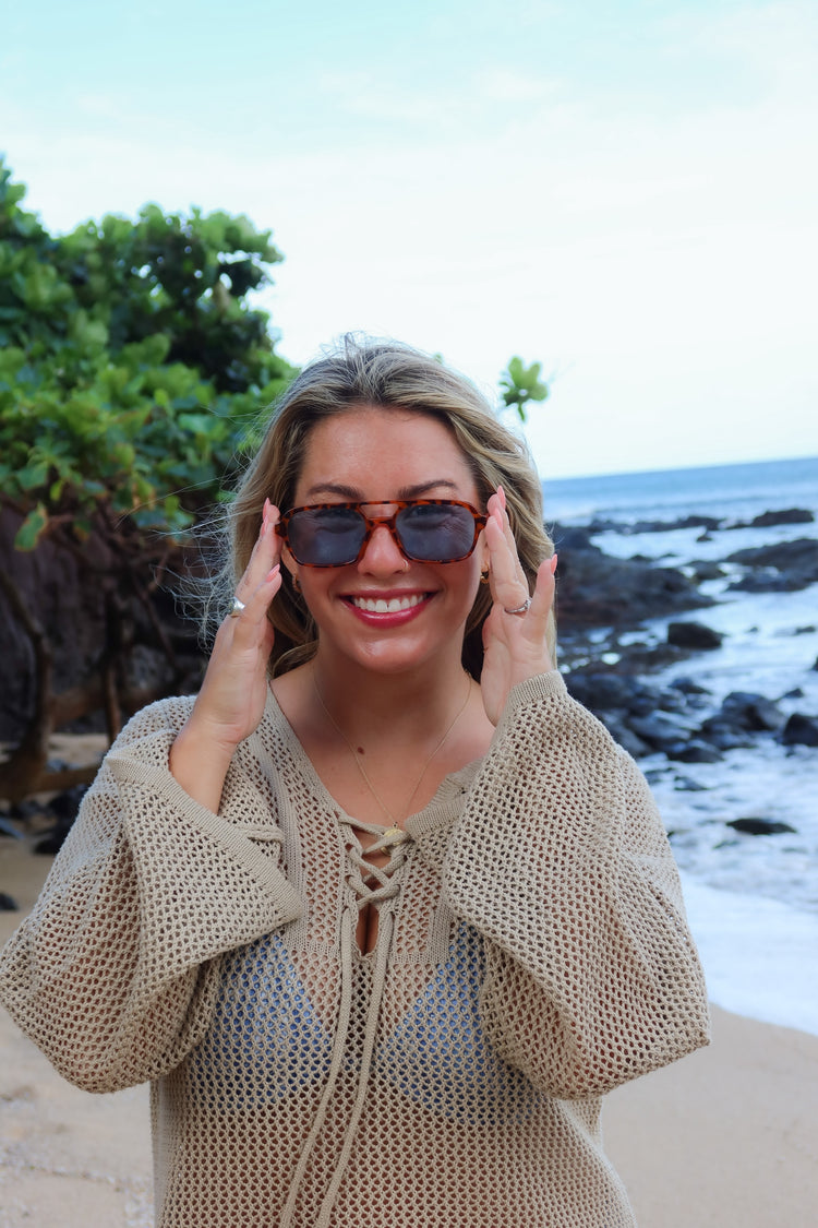 A smiling woman on a beach wearing oversized dark sunglasses and a beige open-knit lace-up top, with rocks and ocean in the background.