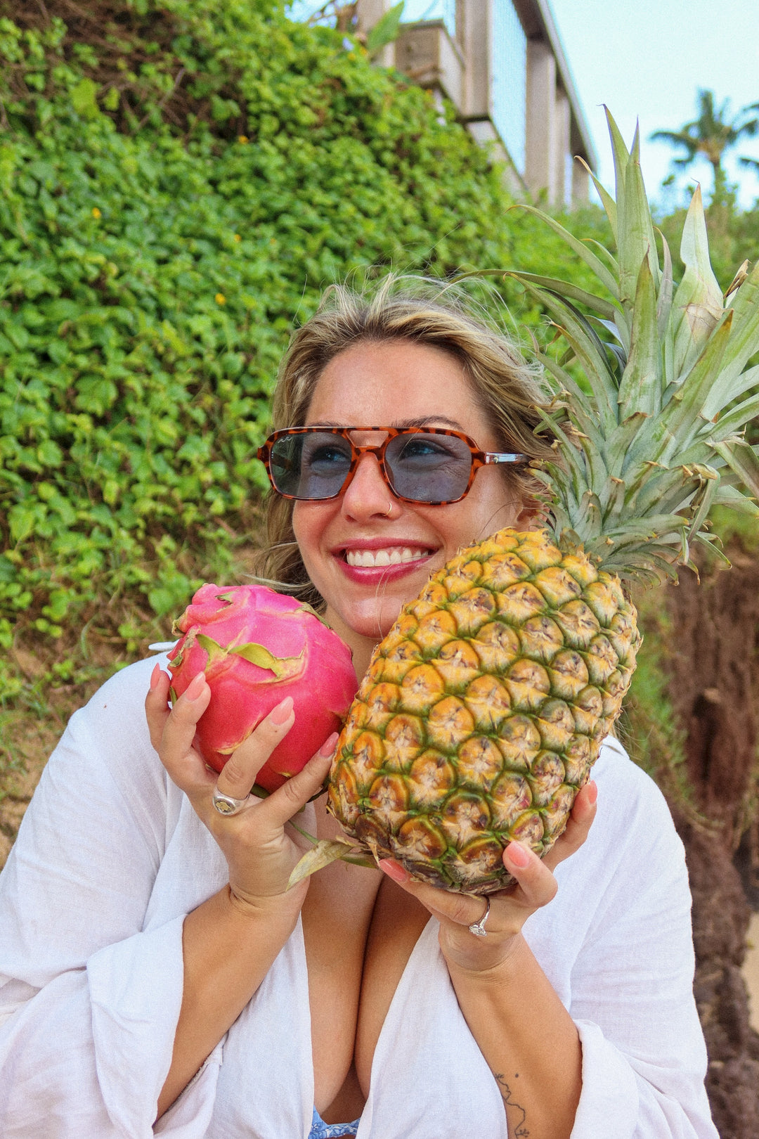 A woman wearing sunglasses and a white open-front top holds a large pineapple and a pink dragon fruit in front of a green leafy background.