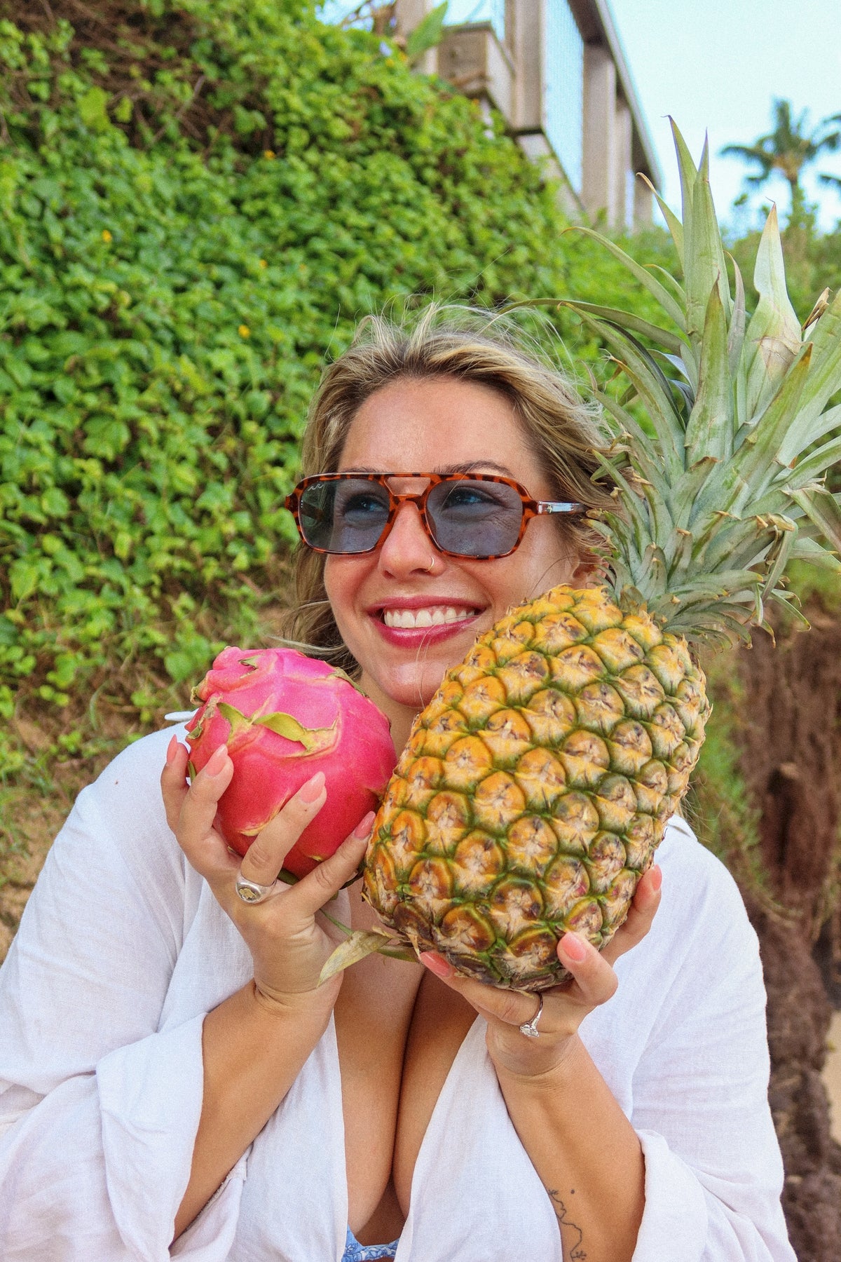 A woman wearing sunglasses and a white open-front top holds a large pineapple and a pink dragon fruit in front of a green leafy background.