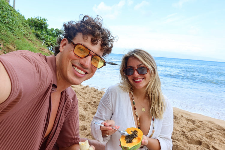 A couple taking a selfie on a sandy beach with the ocean in the background. The man on the left wears a dusty rose-brown shirt and yellow-tinted sunglasses, while the woman on the right wears a white top and sunglasses and is holding a slice of papaya.