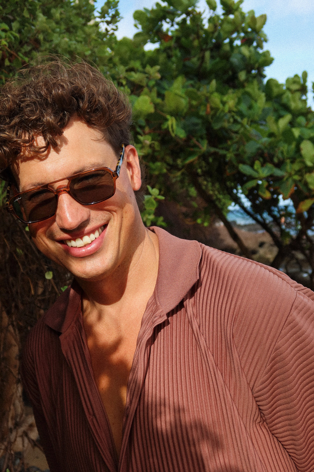 A smiling man outdoors wearing dark aviator-style sunglasses and a brown ribbed open-collar shirt, with green foliage and a blue sky in the background.