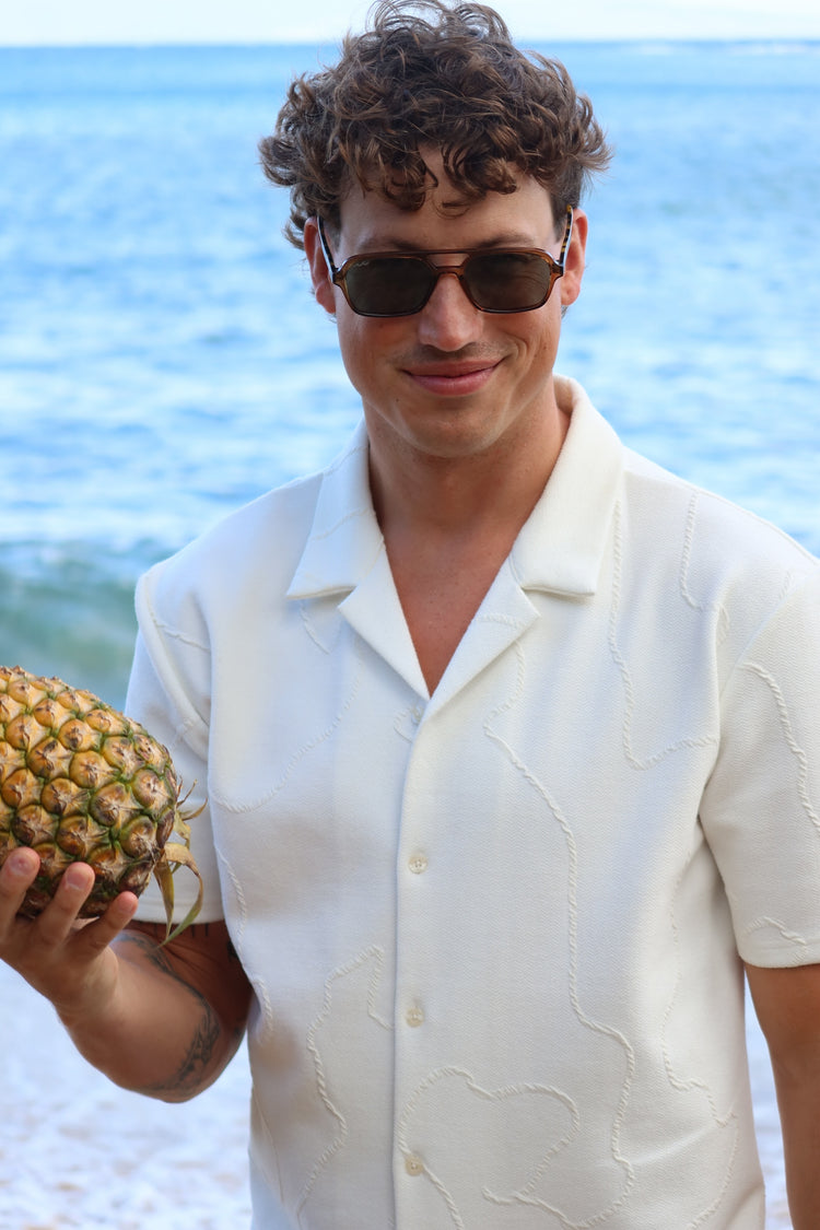 A man wearing a white short-sleeve button-up shirt and sunglasses stands on a beach holding a pineapple, with the ocean in the background.