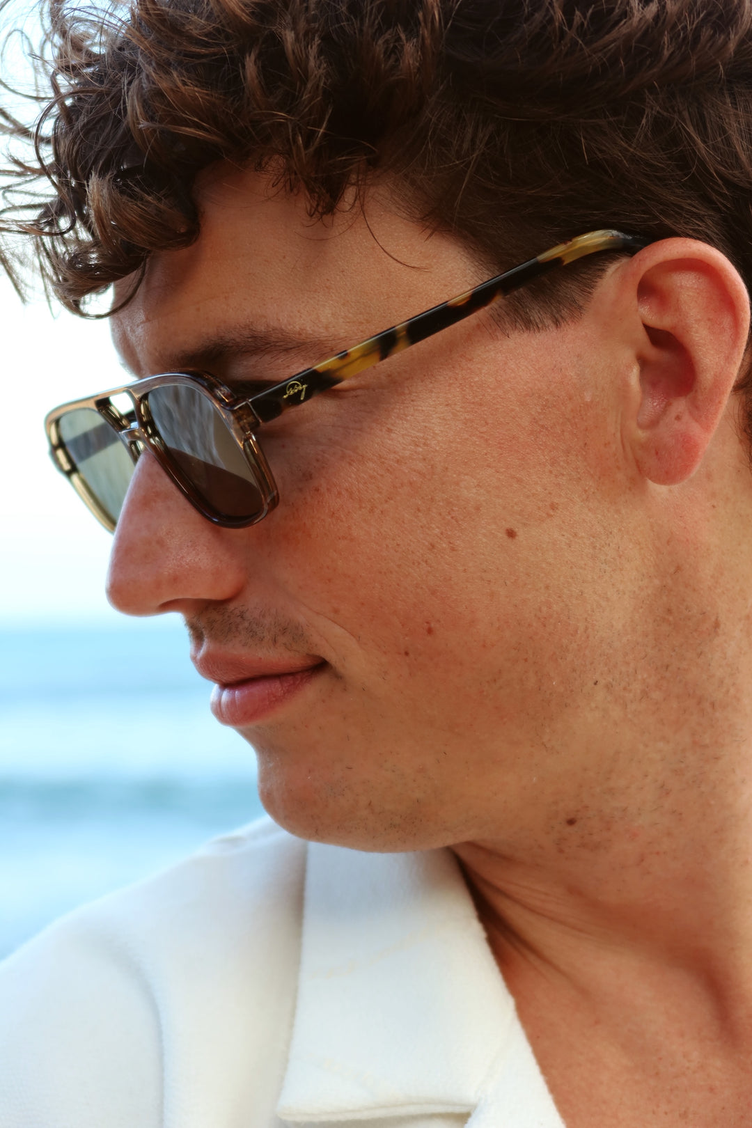 A close-up side profile of a man wearing tortoiseshell aviator sunglasses with gold-toned metal frames and dark lenses, set against a blurred outdoor background.