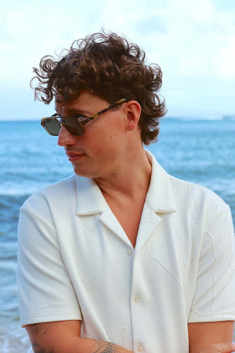 A man with curly brown hair wearing tortoiseshell sunglasses and a white short-sleeve button-up shirt, standing outdoors with the ocean in the background.