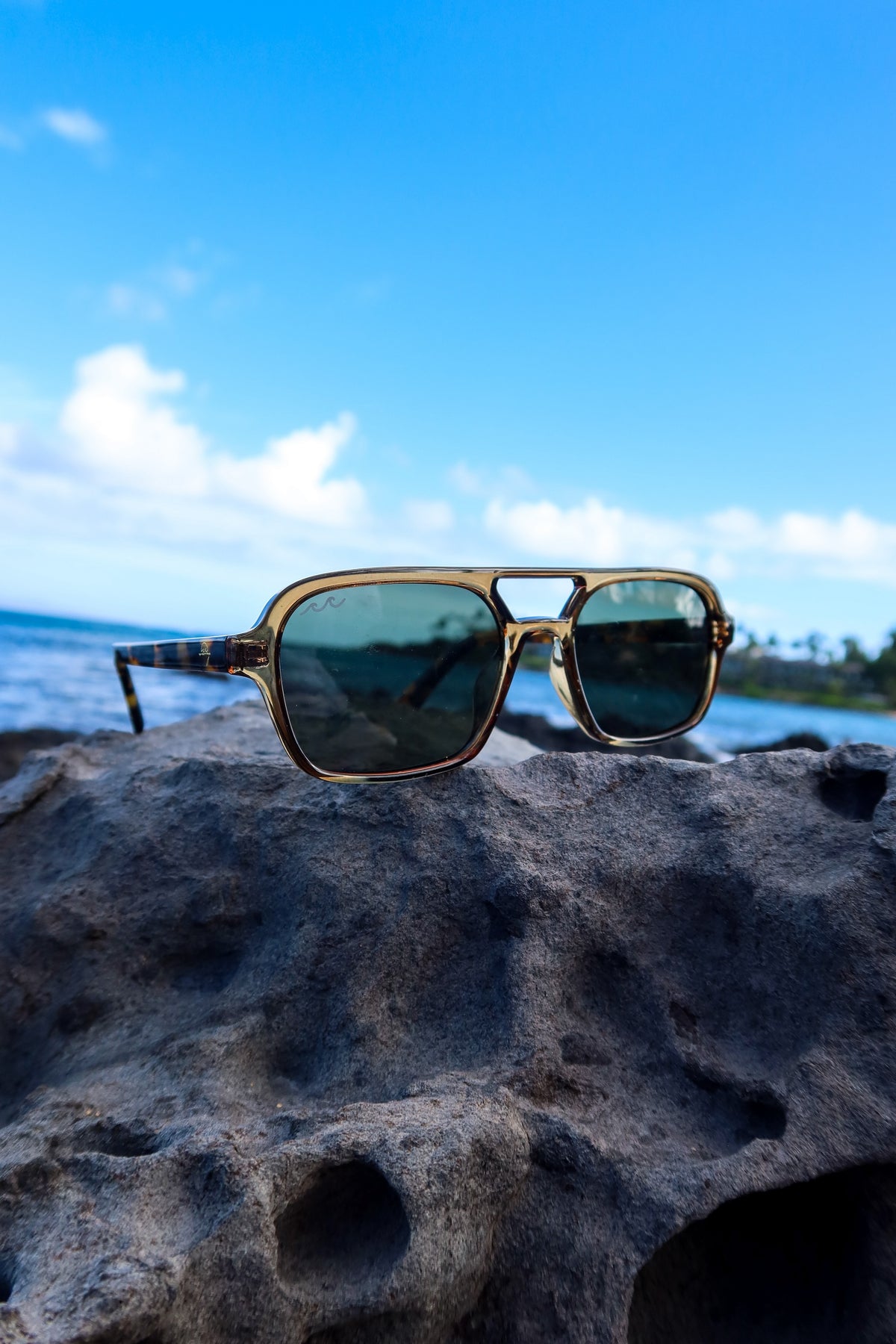 A pair of double-bridge aviator-style sunglasses with a gold-tone metal frame and dark green lenses, resting on a dark volcanic rock with a blue ocean and sky background.