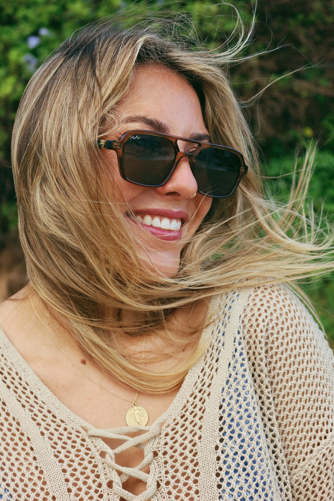 A woman outdoors wearing dark brown square sunglasses with a gold-tone frame and a beige open-knit lace-up top, with her blonde hair blowing in the wind.