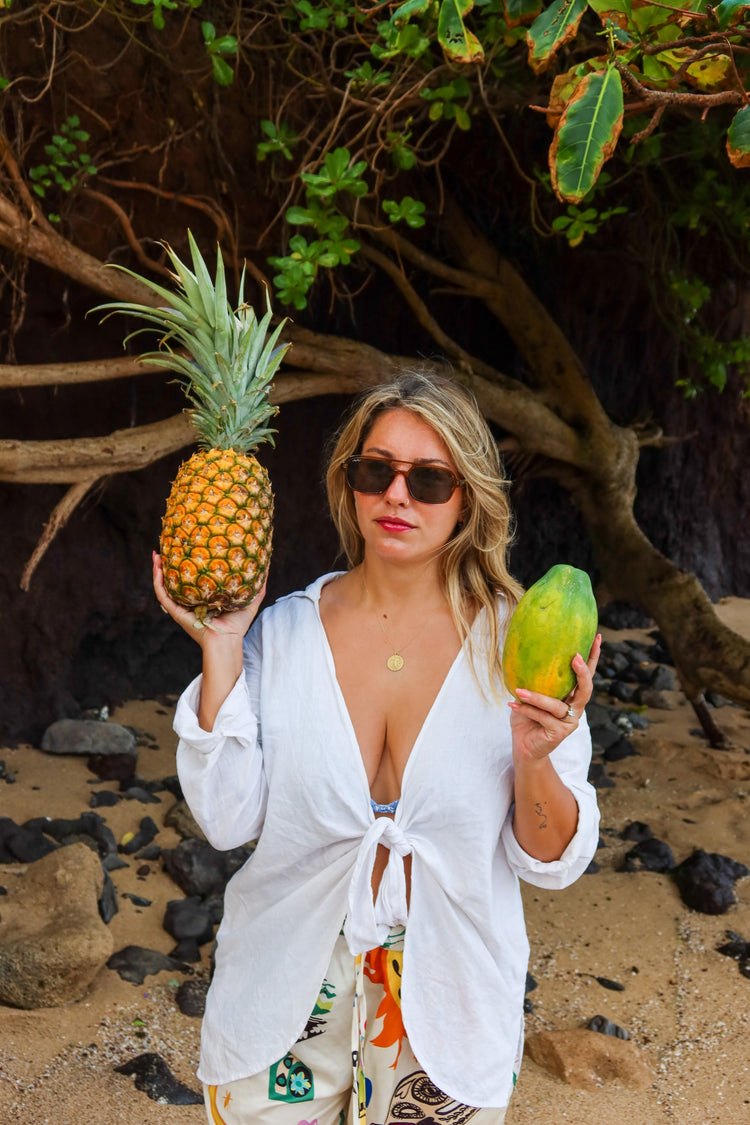 A woman on a beach holds a pineapple in one hand and a green papaya in the other, wearing a white open-front tie-front shirt and patterned bottoms. She is standing in front of a tree and rocks.