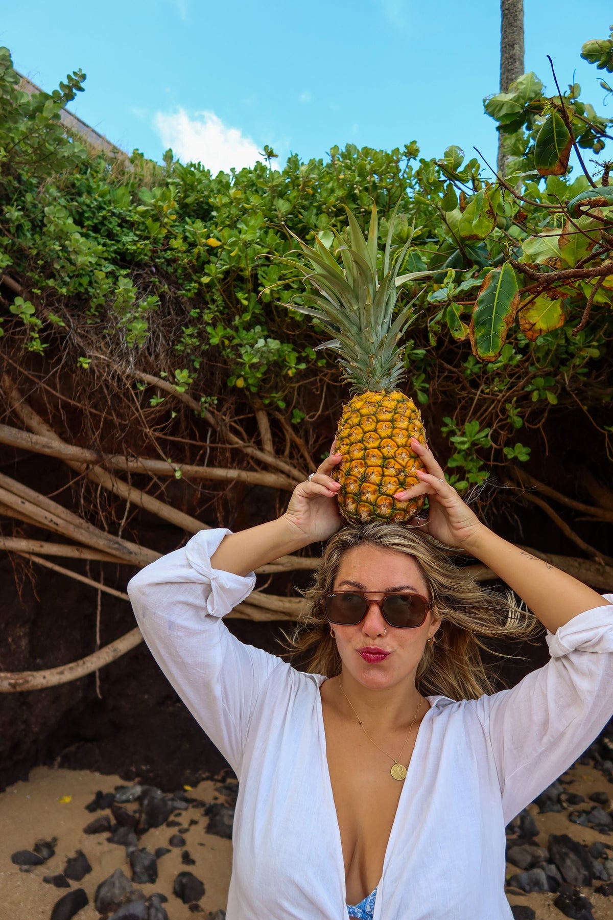 A woman wearing a white button-up shirt and sunglasses balances a whole pineapple on her head outdoors on a sandy beach with green foliage and rocks in the background.
